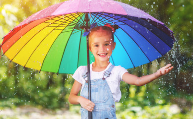 Happy child girl laughs and plays under summer rain with an umbrella.