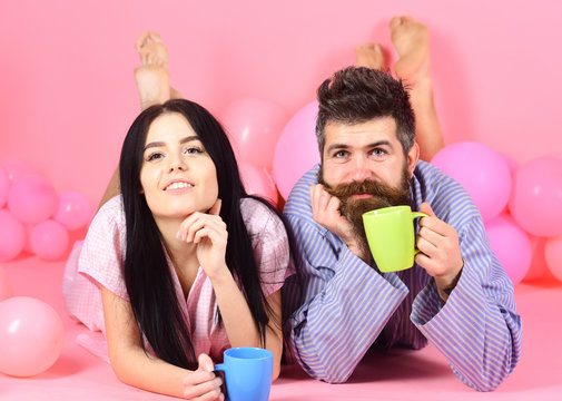 Couple relax in morning with coffee. Man and woman on smiling faces lay, pink background. Weekend morning concept. Man and woman in domestic clothes, pajamas. Couple in love drink coffee in bed.