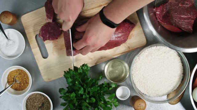 Close up hands of male cook cutting raw meat on wooden cutting board with other ingredients placed around it, top view