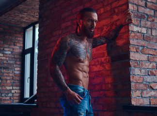 A handsome tattoed shirtless male with a stylish haircut and beard, standing against a brick wall in a room with a loft interior.