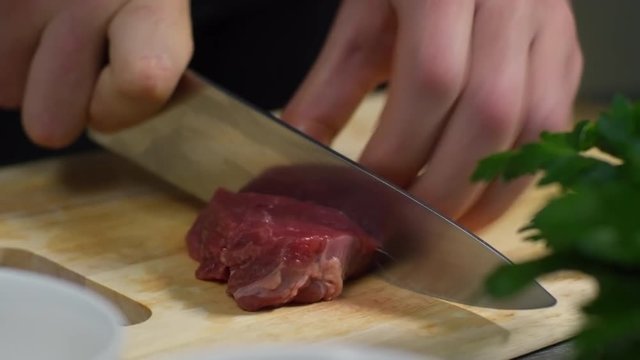 Tracking shot of hands of male cook cutting raw meat on wooden cutting board, close up, tracking right