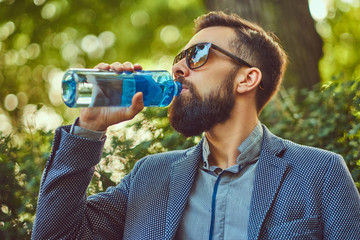 Fototapeta na wymiar Bearded male man drinking cool water outdoors, sitting on a bench in a city park.
