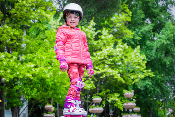 Fototapeta na wymiar A sporty little girl in a helmet and protective gear stands on roller skates in a city park. Beautiful baby in a pink jacket and rollers is on the background of green trees