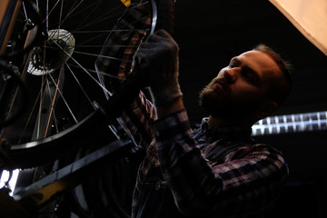 Handsome stylish male wearing a flannel shirt and jeans coverall, working with a bicycle wheel in a repair shop.