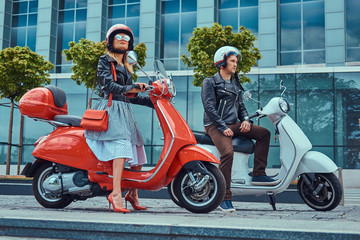 Attractive romantic couple, a handsome man and sexy female, sitting on retro Italian scooters against a skyscraper.