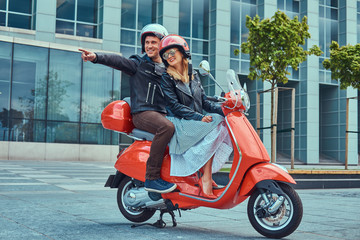 Fototapeta na wymiar Attractive happy couple, a handsome man and sexy female riding together on a red retro scooter in a city.