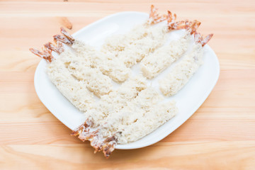 Close-up of raw tempura shrimps in a breaded lay on a white plate that stands on a wooden table. Marine semi-finished products for home cooking