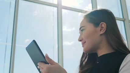 Young female passenger at the airport, using her tablet computer while waiting for her flight, smiling, sunnu day, terminal raiyway station bus transportation, business woman student