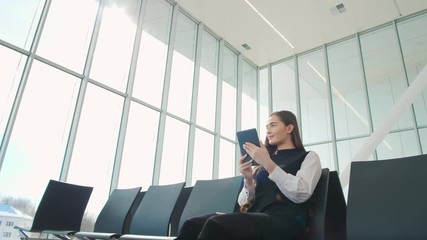 Young female passenger at the airport, using her tablet computer while waiting for her flight, smiling, sunnu day, terminal raiyway station bus transportation, business woman student