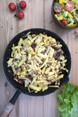 Fried potatoes in an old frying pan, tomato lettuce leaves and fresh vegetable salad on a wooden background.