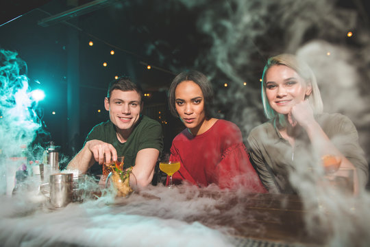 Portrait of happy girls and glad male telling together while sitting in bar. They situating in smoke. Positive friends drinking glasses of alcohol beverage concept