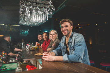 Portrait of cheerful men and outgoing girls looking at camera while sitting at counter in bar. Smiling comrades tasting alcohol concept