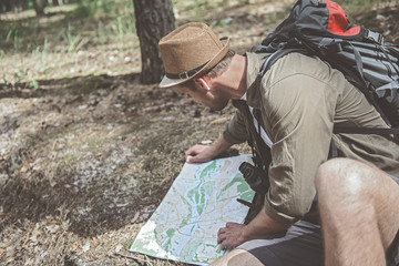 Looking for directions. Top view of young traveler with backpack is sitting on the ground in pine forest and looking at map. He is checking all ways. Copy space in the left side