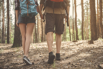 Spend time together. Low angle close-up of legs of pleasant young tourists are holding hands while going across wood with backpacks. Back view
