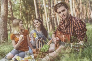 Naklejka na ściany i meble Rejoicing friends taking seat on rug with food. Calm man with guitar in hands looking at camera. Two girls drinking from disposable cups and laughing