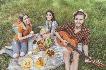 Portrait of delighted people arranging picnic outside. Guy sitting with guitar and girls resting with beverages. They looking at camera with pleasure