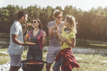 Laughing campers standing near brazier with alcohol in hands. Men feeding women with barbecue. River on background