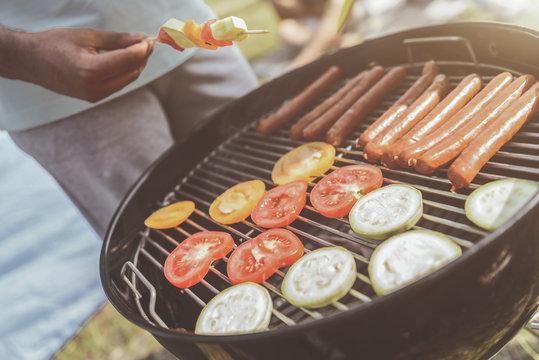 male hands roasting tasty food on grill. Focus on grate with vegetable and sausages on it. Close up