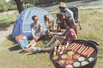 Focus on sausages and vegetables on grill. Men and women sitting with guitar and beer on background