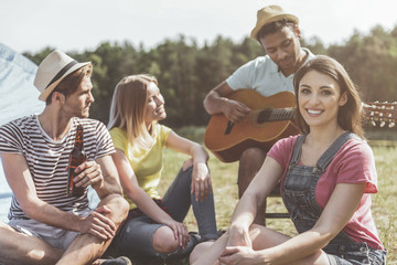 Calm people sitting on grass. Concentrated man performing song on guitar while companions listening and smiling. Focus on woman looking at camera with joy