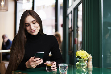 Young woman with long hair texting by mobile phone, smiling, drinking coffee having rest in cafe near window
