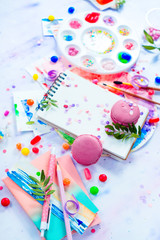 Pink macarons in a colorful party activity concept with confetti, artist tools, brushes, palette and sketchbook with blank pages. Creative celebration flat lay with copy space.