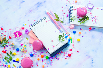 Planning a party concept with confetti, pink macarons, candies and an open notepad with blank pages. Creative celebration flat lay with copy space.