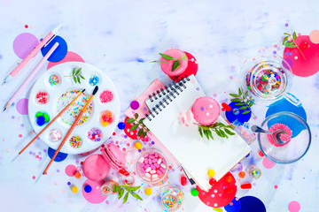 Fototapeta na wymiar Sweettooth artist tools creative party concept. Pink macarons in a colorful party activity concept with confetti, artist tools, brushes, palette and sketchbook with blank pages.