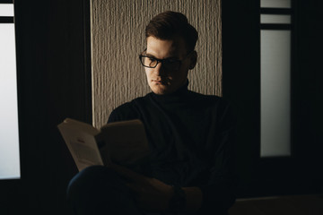 Handsome young man in eyeglasses reading book at home.