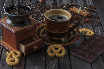 on a dark wooden table a coffee grinder, a cup of coffee, chocolate and pretzels