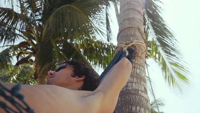 Young handsome man lies on hammock under palm trree relaxing on beautiful exotic beach. vacation concept. slow motion. 1920x1080