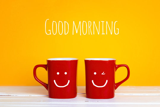Two red coffee mugs with a smiling faces on a yellow background with the phrase Good morning.