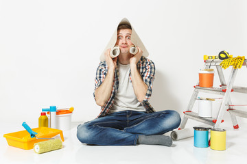 Fun young male sitting on floor with roll of wallpaper above head, instruments for renovation apartment room isolated on white background. Gluing accessories, painting tools. Repair home concept.