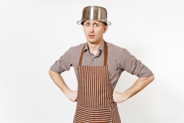 Fun man in striped apron with silver stainless glossy aluminium empty stewpan, pan or pot on head isolated on white background. Male housekeeper or houseworker. Kitchenware, dishes, cuisine concept.