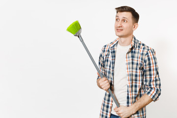 Young tired upset housekeeper man in checkered shirt holding and sweeping with green broom isolated on white background. Male doing house chores. Copy space for advertisement. Cleanliness concept.