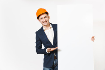 Young smiling businessman in dark suit, protective hardhat holding big white empty blank material with place for text or image and pointing hand aside on copy space isolated on white background.