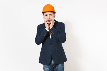 Young handsome concerned businessman in dark suit, protective construction orange helmet keeping hands near face isolated on white background. Male worker for advertisement. Business, working concept.