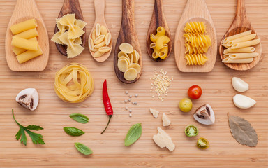 Italian food concept .Various kind of pasta in wooden spoons with ingredients sweet basil ,tomato ,garlic ,parsley ,bay leaves ,pepper ,champignon and parmesan cheese on bamboo cutting board flat lay.