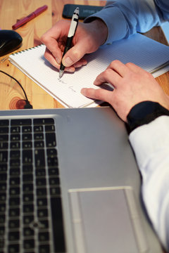 Man hands wearing light blue striped shirt work with laptop and write with pen on paper on old natural wooden desk at work-space in home. Working from home concept at shallow depth of field.