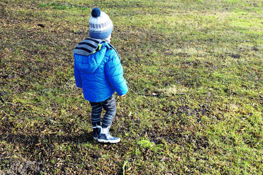 Two years old boy wearing blue hooded outdoor jacket, knitted hat and rubber rain boots walking around green brown spring grass in yard. Horizontal image with copy space for text.
