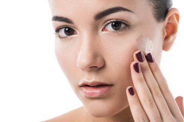 attractive girl applying foundation cream on cheek and looking at camera isolated on white