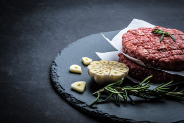 Raw burger beef patty with rosemary and garlic on dark cooking background