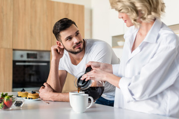 young woman pouring coffee and looking at handsome boyfriend at morning in kitchen