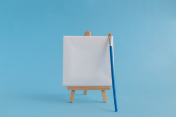 Blank art board canvas with wooden stand miniature and paintbrush on plain blue background. Art equipment minimalistic concept.