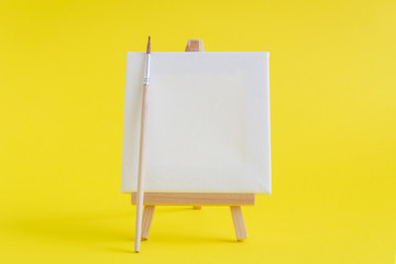 Blank art board canvas with wooden stand miniature and paintbrush on plain yellow background. Art equipment minimalistic concept.