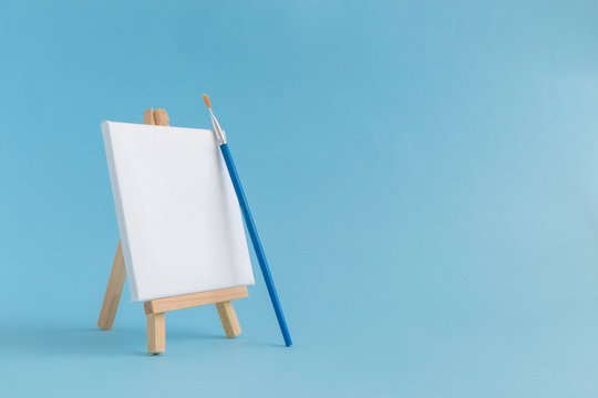 Blank art board canvas with wooden stand miniature and paintbrush on plain blue background. Art equipment minimalistic concept.
