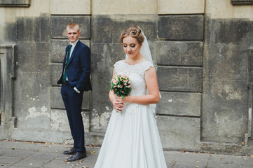 Wedding blonde couple is walking in the old city. Groom is standing near the ancient stone walls. Bride in lace satin dress is holding pink rose bouquet. Sunny love story in the medieval town.