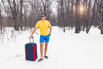 Travelling from winter to summer. Young cute guy in sunglasses, flip-flops, t-shirt and shorts with a tourist suitcase goes through the forest in the snow. Dream of vacation, sea, warm exotic country.