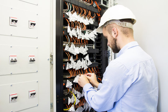Engineer performs adjustment of computer network cabinet with controllers, switches and optical cables. A worker in white helmet connects fiber to telecommunications equipment switches in data center