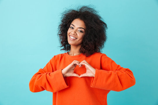 Adorable american woman in colorful shirt looking on camera and gesturing heart shape with hands, isolated over blue background